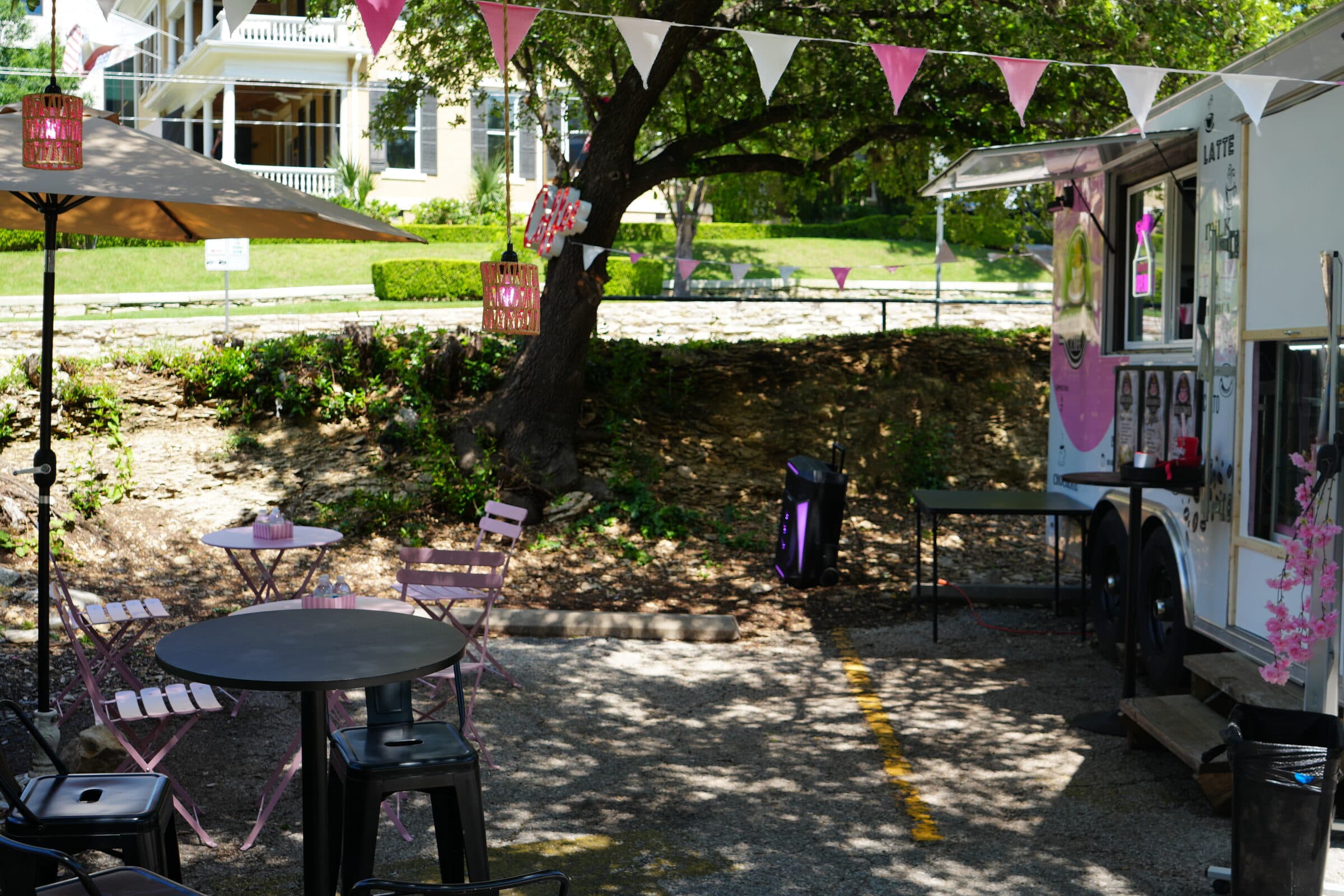 Coffee truck patio on the ANW lot with a pink umbrella, café tables, and bunting under a tree